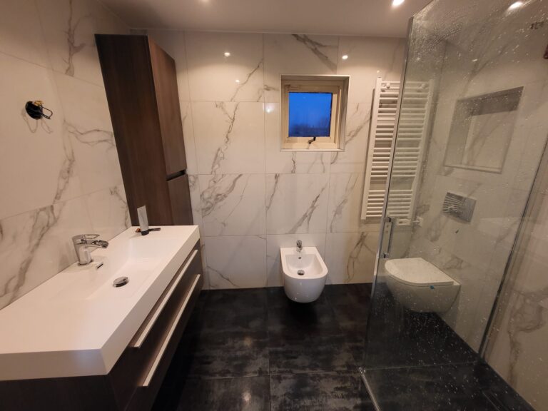 Renovated bathroom with white marble-look wall tiles, dark floor, wooden vanity unit and walk-in glass shower