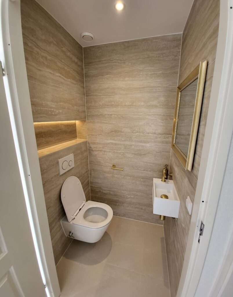 Renovated toilet with travertine-look tiles, gold-framed round mirror and wall-hung toilet