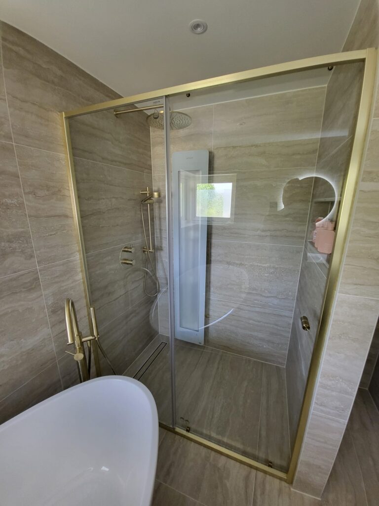 Walk-in shower with gold-framed glass enclosure and travertine-look tiles beside a freestanding bath