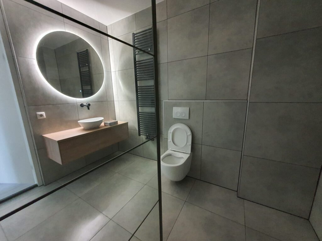 Renovated bathroom with large grey tiles, round LED mirror, wooden vanity and walk-in glass shower