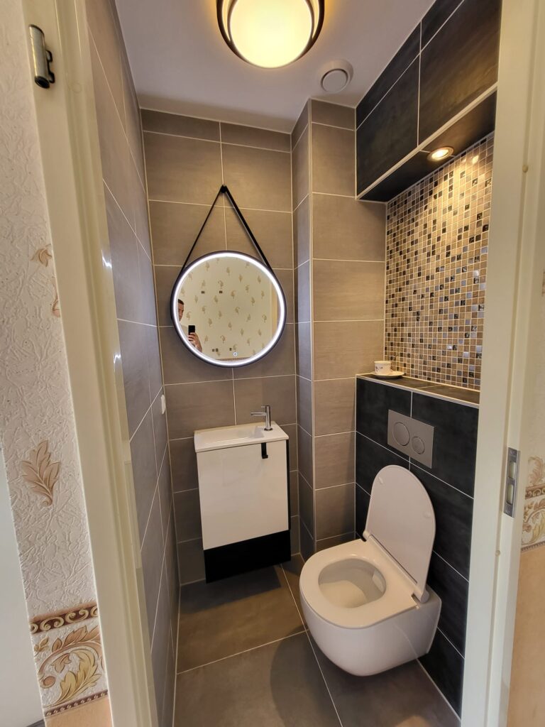 Renovated toilet with grey tiles, mosaic accent strip, round mirror and wall-hung toilet